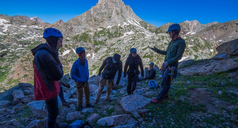 A group of people listen to an instructor speak. They are all wearing helmets, and there is a rocky mountain behind them.